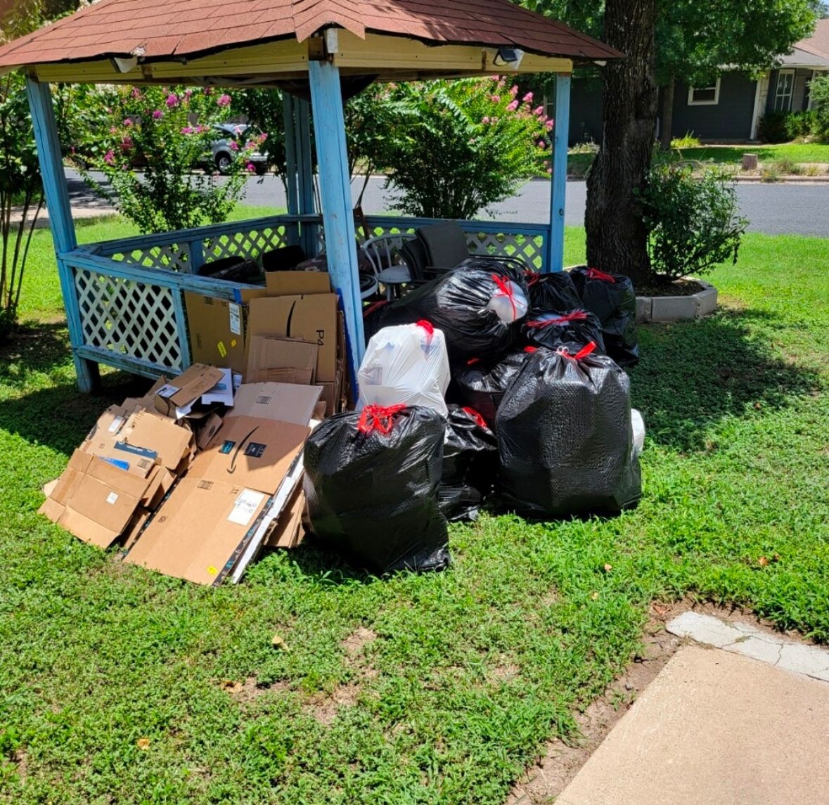 Pile of black trash bags and cardboard boxes under a blue wooden gazebo on a grassy lawn.